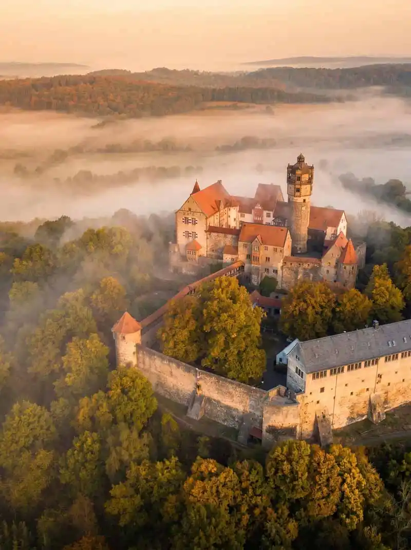 Eine neblige Luftaufnahme von Ronneburg, einer mittelalterlichen Burg mit roten Dächern und einem zentralen Turm, umgeben von dichtem Wald und alten Steinmauern an einem nebligen Morgen, in der Nähe des historischen Gasthauses, das für seine hervorragende Küche bekannt ist.